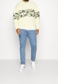Sweat-shirt jaune clair avec un motif de palmier sur la poitrine. Associé à un jean bleu et des baskets blanches. Coupe décontractée et style casual.