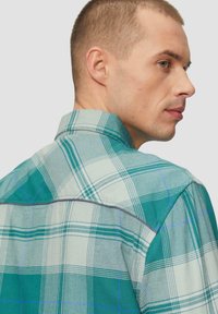 Koszula w kratę w odcieniach turkusu, bieli i niebieskiego. Posiada klasyczny kołnierzyk, krótkie rękawy i szczegółowe przeszycia wzdłuż pleców.