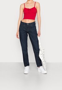 Röd kort topp med justerbara remmar, i kombination med mörkblå raka jeans. Modellen håller en vit väska med svarta mönster. Vita sneakers.