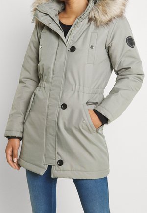 Personne portant un manteau d'hiver gris clair avec des boutons noirs, une capuche bordée de fourrure et les mains dans les poches latérales, associée à un jean bleu.