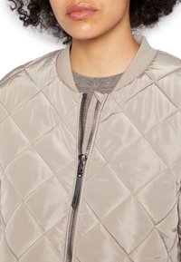 Beige kviltad bomberjacka med en slät, glänsande yta, ribbad krage och full dragkedja. Anmärkningsvärd svart dragkedjedetalj.