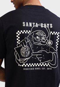 Μαύρο βαμβακερό μπλουζάκι με λευκό σχέδιο του Άγιου Βασίλη που κρατάει μια κάρτα με την ένδειξη "Εγκεκριμένο", με καρό διακοσμητικά και το κείμενο "SANTA SAYS."
