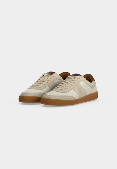 Paire de baskets basses beige avec semelles en caoutchouc naturel, fermeture à lacets à l’avant et languettes marron au talon, présentée sur fond blanc.