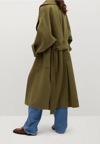 Oliivinvihreä villatakki, jossa on vyö, leveät hihat ja kaksi sivutaskua. Päällä vaaleansiniset farkut ja ruskeat kengät, joissa on kultainen koriste.