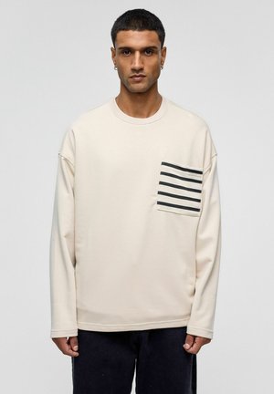 Lichtbeige sweatshirt met lange mouwen en een zwart-wit gestreepte zak op de linkerborst. Zachte textuur, comfortabel model, ronde halslijn.