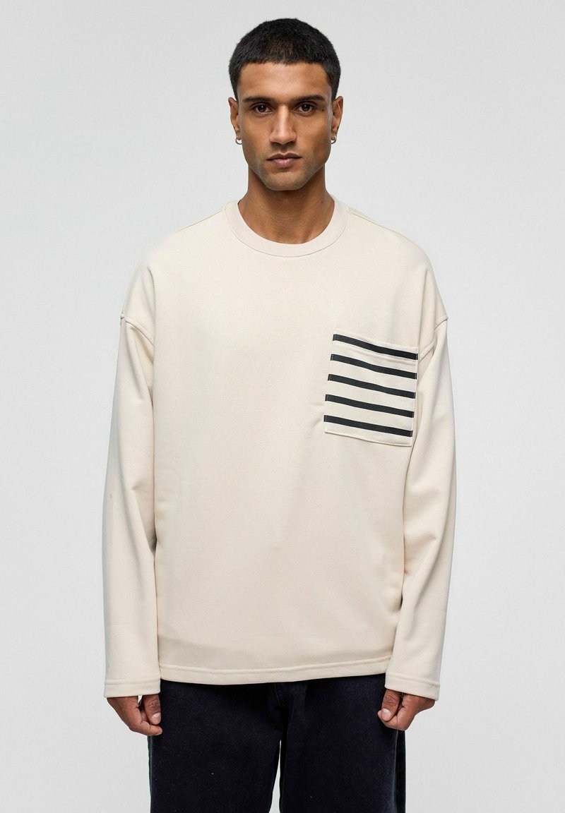 Lichtbeige sweatshirt met lange mouwen en een zwart-wit gestreepte zak op de linkerborst. Zachte textuur, comfortabel model, ronde halslijn.