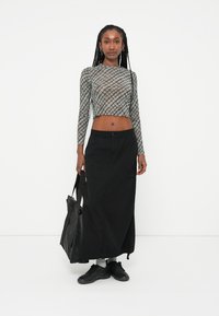 Saia longa preta com bolsos de carga, combinada com um top cropped cinza claro e padrão, justo e transparente. Modelo usa uma bolsa tote preta e calça ténis pretos.
