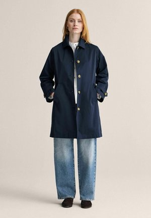 Punahiuksinen nainen pukeutuneena laivastonsiniseen napitettuun takkiin, vaaleanpesuisiin leveälahkeisiin farkkuihin ja tummiin mokkasiinikengiin, seisoo kädet takin taskuissa.