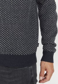 Striktrøje med et mørkeblått og lysegråt mønster, ribbede manchetter og kant, med en struktureret finish og synlig mærkeetiket på siden.