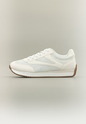 Witte lage sneaker met mesh en leren panelen, witte veters en een bruine rubberen zool, zijaanzicht op een beige achtergrond.