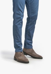 Mann in blauen Slim-Fit-Hosen und grauen Wildleder-Schnürschuhen, der vor einem weißen Hintergrund steht.