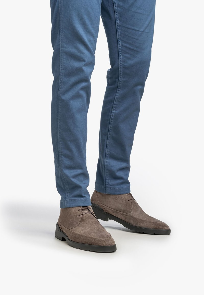 Mann in blauen Slim-Fit-Hosen und grauen Wildleder-Schnürschuhen, der vor einem weißen Hintergrund steht.