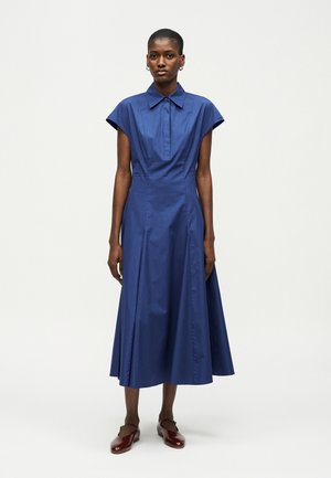 Γυναίκα με κοντά μαλλιά που φοράει μπλε midi φόρεμα χωρίς μανίκια με γιακά και εμπρόσθιο πόδι, συνδυασμένο με σκουρό κόκκινα επίπεδα παπούτσια, στέκεται σε λευκό φόντο.