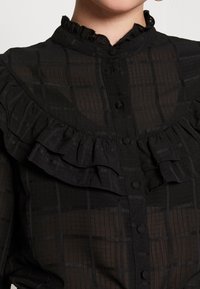 Sort, gennemsigtig bluse med en flæset halsudskæring, knaplukning og et ternet mønster med varierende teksturer.