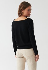 Maglione nero lavorato a maglia con ampio scollo a barca e maniche lunghe, caratterizzato da un orlo a coste. Il modello lo abbina a jeans beige.