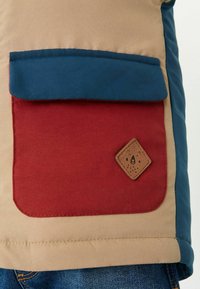 Veste beige avec une poche avant rouge et un rabat bleu, ornée d'un petit patch marron avec un visage d'ours dans le coin de la poche, portée avec un jean bleu.