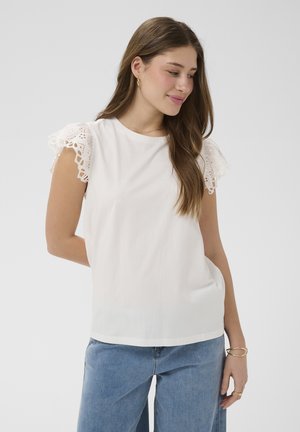 Jeune femme aux longs cheveux bruns portant un haut blanc avec des manches courtes en dentelle et un jean bleu clair, debout devant un fond clair uni.