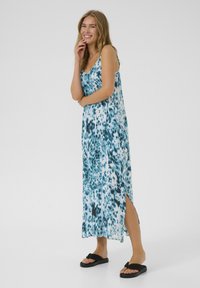 Robe maxi bleu et blanche avec un motif de feuilles, bretelles spaghetti, fentes latérales, confectionnée en tissu léger, associée à des sandales noires.