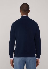 Granatowy sweter z dzianiny o prążkowanej strukturze, z kołnierzem stojącym i długimi rękawami, w zestawieniu z jasnoniebieskimi spodniami i mankietem w paski.