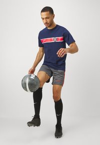 Tamsiai mėlyna trumpomis rankovėmis marškinėliai su raudona juostele ir logotipu, pilki šortai su raudonais akcentais, juodos iki kelių kojinės ir juodi futbolo batai.