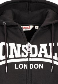 Zwarte ritsvest gemaakt van katoen met witte tekst en logo "LONSDALE LONDON". Voorzien van trekkoorden en een merkrits. Maat M.