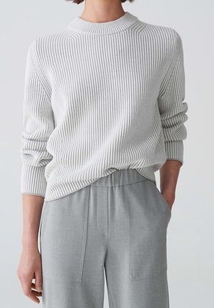Personne portant un pull en maille côtelée blanc à manches longues et un pantalon taille haute gris clair avec poches, debout devant un fond uni.