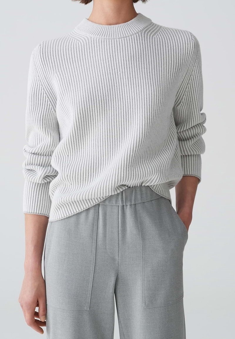 Personne portant un pull en maille côtelée blanc à manches longues et un pantalon taille haute gris clair avec poches, debout devant un fond uni.