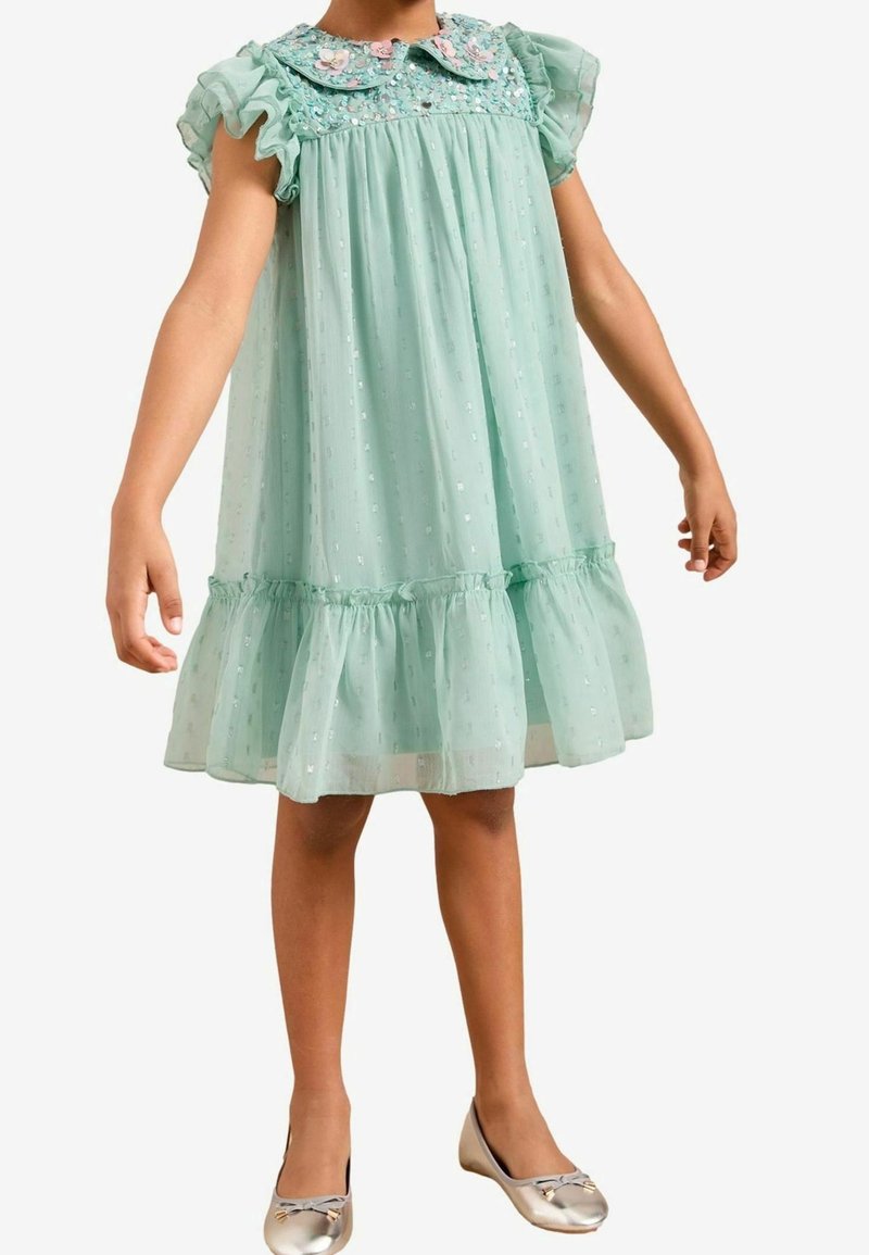 Miętowa sukienka z krótkimi, falbankowymi rękawami i kołnierzykiem z kwiatowym haftem. Przezroczysta tkanina z marszczonym, warstwowym dołem. Srebrne baleriny.