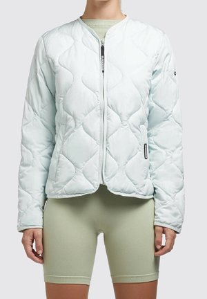 Nainen, joka pukeutunut vaaleansiniseen tikattuun vetoketjutakkiin ja vaaleanvihreisiin istuviin pyöräilyshortseihin, seisoo yksiväristä taustaa vasten.