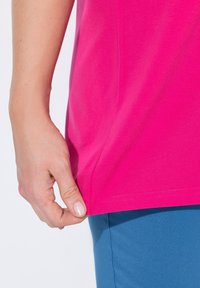 Camiseta de algodón fucsia con manga corta, que presenta un dobladillo recto y una abertura lateral, usada sobre pantalones ajustados azules. Sostenida a mano en el dobladillo.