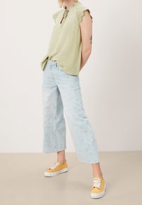 Blusa verde claro con acentos de encaje y cuello de lazo; combinada con vaqueros de pierna ancha en azul desvaído con patrones florales; zapatillas amarillas.