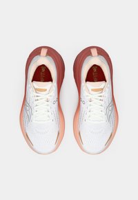 Baskets blanches avec une tige respirante, accents roses, semelle épaisse et motif texturé. Elles comportent des lacets et un tirant à l'arrière.