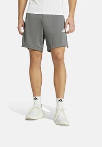 Pantalones cortos deportivos grises con una textura suave, que presentan una cintura elástica y el logo de Adidas en la pierna izquierda. Combinados con zapatos y calcetines blancos.