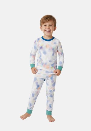 Lachende jonge jongen die blootsvoets staat en een wit pyjamapak draagt met kleurrijke aquarelpatronen en groene boorden aan mouwen en broek.