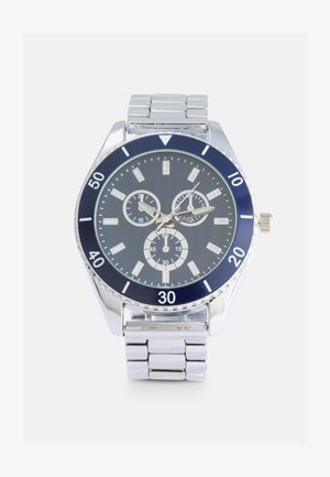 Silberne Edelstahl-Armbanduhr mit einem navyblauen Lünette, drei Zifferblättern und silbernen Stundenmarkierungen auf einem strukturierten dunkelblauen Hintergrund.