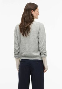 Svetlo sivi pleteni pulover z zvitimi rokavi, kombiniran z mornarsko modrimi hlačami, ki imajo dva zadnja žepa in gumbne okraske. Vidna rebrasta spodnja obroba.