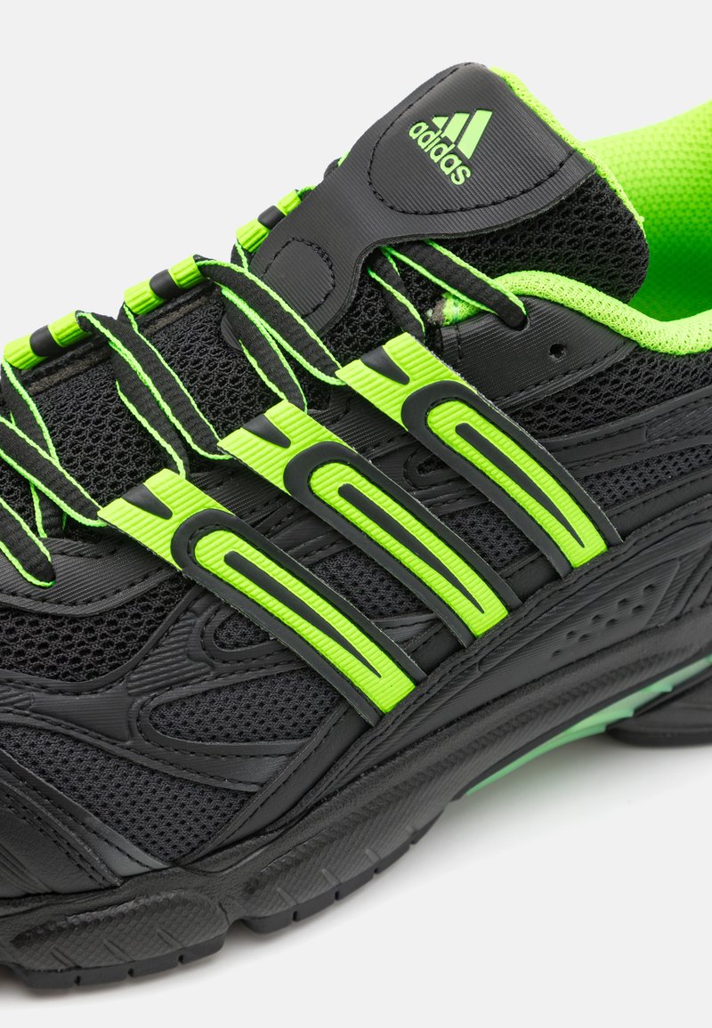 Zapatilla deportiva negra con acentos verdes; cuenta con una parte superior de malla, un sistema de cordones gruesos y una suela texturizada para agarre. Diseño notable de tres rayas.