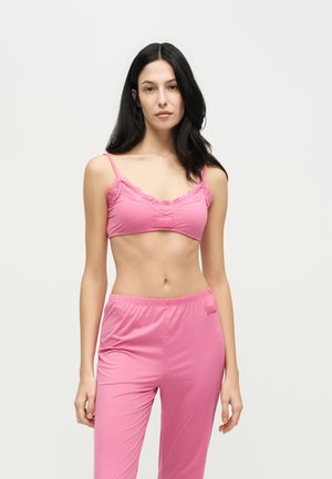 Un modèle aux longs cheveux foncés pose sur un fond uni, portant un caraco rose bordé de dentelle et un pantalon assorti.