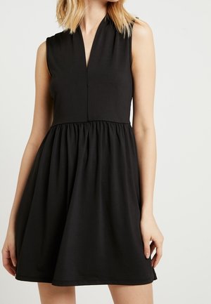 Sleeveless black dress with V-neckline and fitted bodice, flared gathered skirt, worn by standing person with blonde hair.