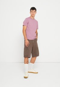 Lyse lilla kortærmet t-shirt sat sammen med brune cargo shorts. Model iført hvide sokker og beige sneakers, står foran en hvid baggrund.