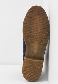 Semelle d'une chaussure foncée avec une semelle en caoutchouc marron texturé, numéro de taille visible et logo circulaire, présentée sur fond blanc.