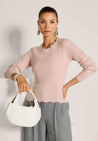 Frau, die einen hellrosa gerippten Pullover, graue Hosen und silberne Creolen trägt, während sie eine weiße Handtasche mit einem geschwungenen Henkel hält.