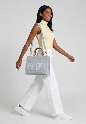 Mujer caminando, con una blusa sin mangas amarilla y pantalones blancos, llevando un bolso gris con el texto "SUMMER VIBES" y diseños de corazones.