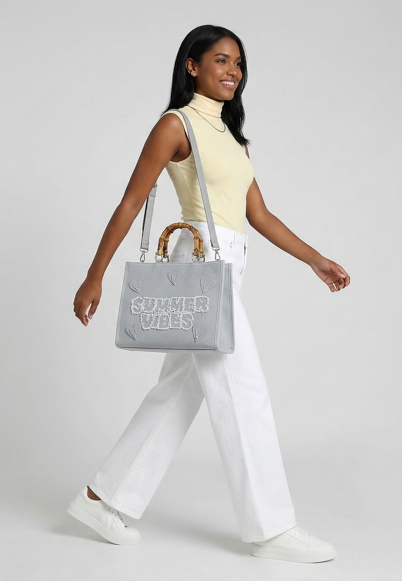 Frau geht, trägt ein gelbes ärmelloses Oberteil und weiße Hose, trägt eine graue Handtasche mit dem Schriftzug "SUMMER VIBES" und Herzmotiven.