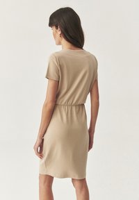 Abito beige a maniche corte con vita arricciata, caratterizzato da un collo rotondo e una texture liscia, che termina appena sopra il ginocchio. Nessun motivo visibile.