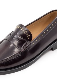 Mocassino in pelle bordeaux con borchie dorate sulla parte superiore, caratterizzato da un design cucito e una suola in gomma nera bassa. Texture liscia e lucida.