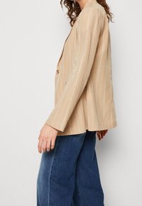 Person klädd i en beige rutig blazer och blå vida jeans, sedd från sidan mot en enfärgad ljus bakgrund.