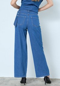 Pantaloni albastri din denim cu croială largă și textură cu dungi fine, cinci buzunare și clapetă decorativă laterală. Finisați cu tiv neprelucrat și purtați cu pantofi cu toc negri.