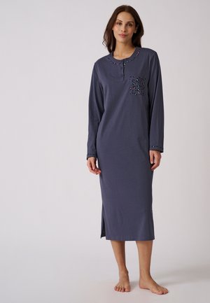 Vrouw staat op blote voeten en draagt een donkerblauwe nachtjapon met lange mouwen en tot aan de enkels, met een patroon op de kraag en borstzak.