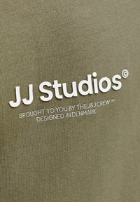 Tissu vert olive avec tricot texturé ; présente un logo texte blanc "JJ Studios" et un marquage supplémentaire en plus petit.